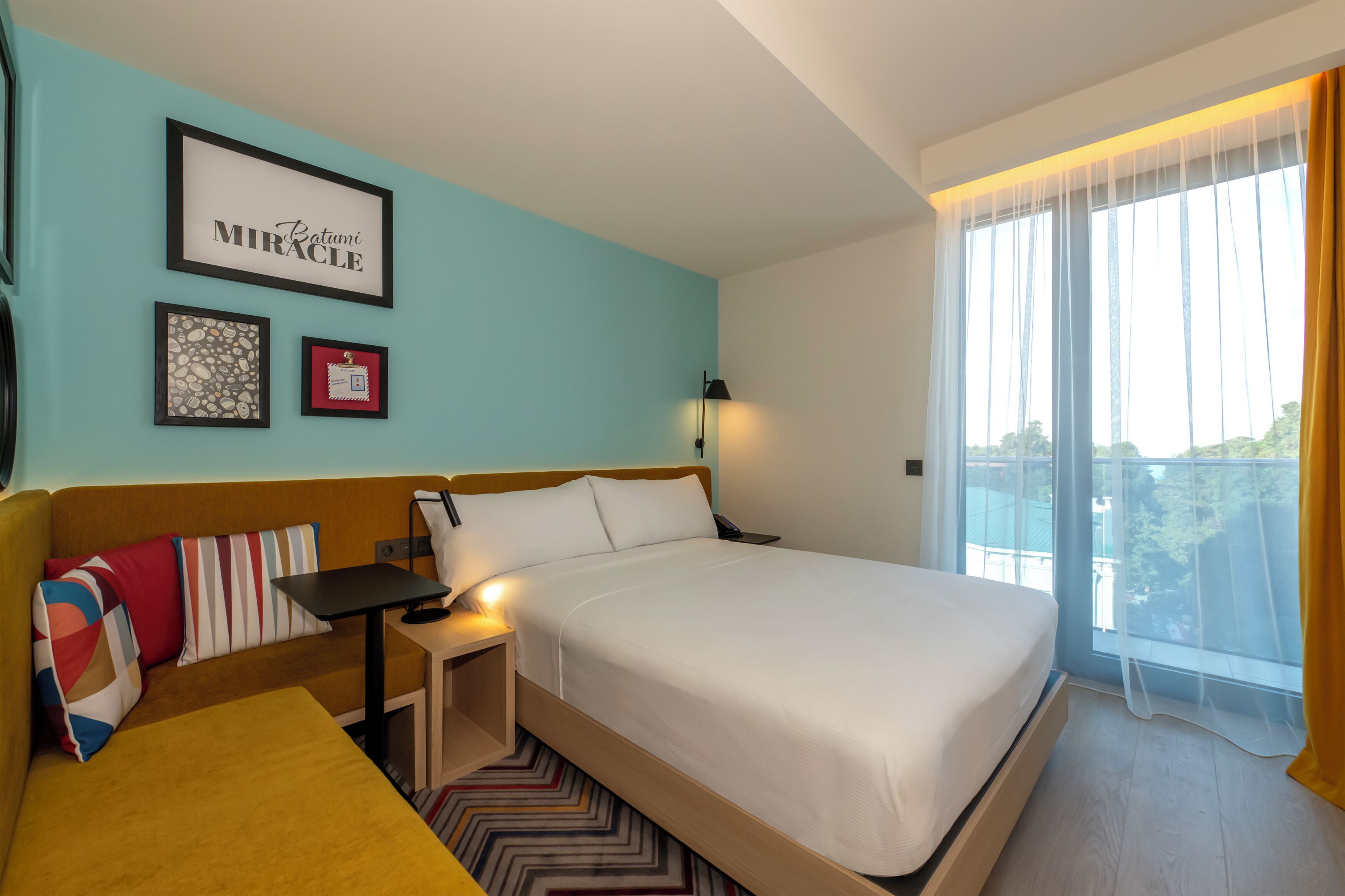 A hotel room with a bed, a sofa, a small table, a lamp, and a window with curtains.