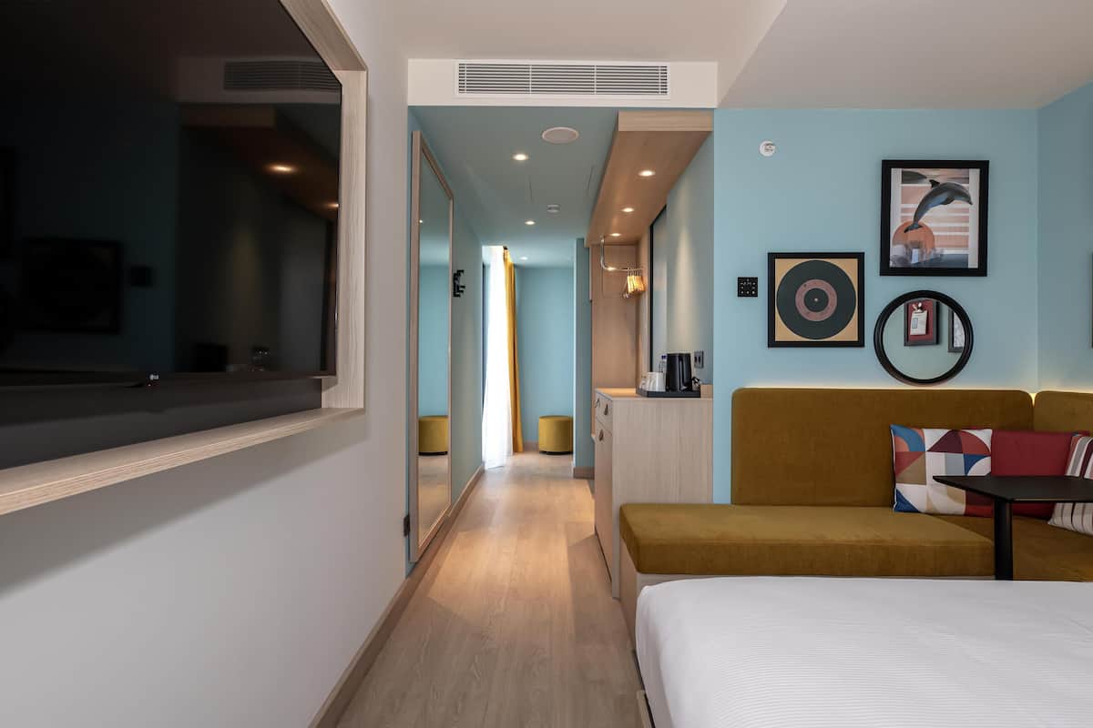 A hotel room with a bed, a sofa, a small table, a mirror, and a wall with framed pictures.