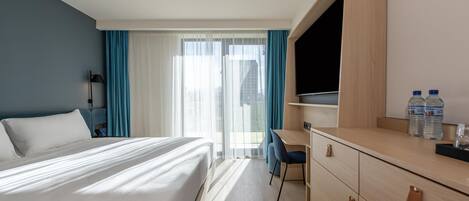 A hotel room with a bed, a desk, a chair, a TV, and a view of the city through the window.