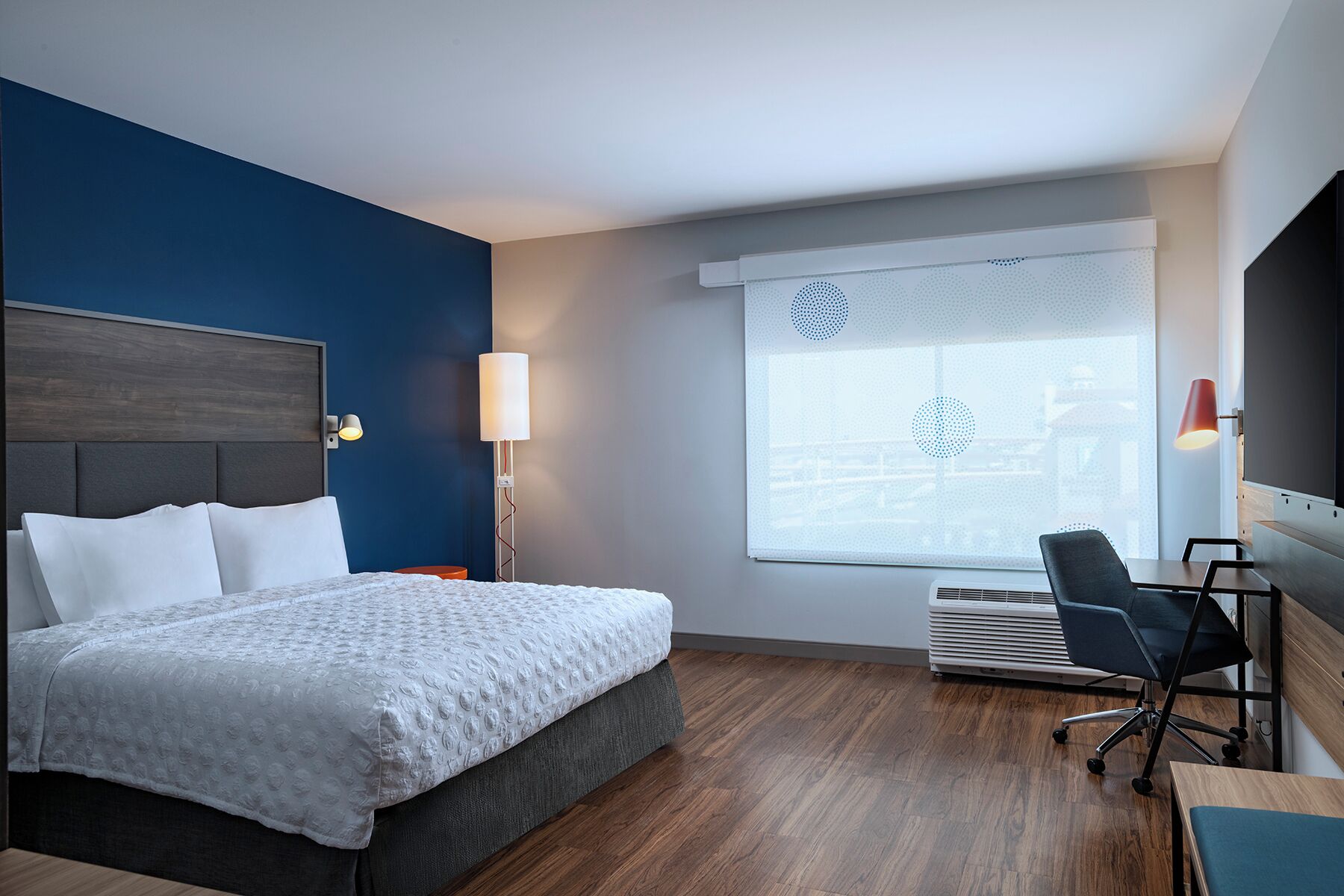 Ein Hotelzimmer mit Bett, Schreibtisch, Stuhl und großem Fenster mit Jalousien.
