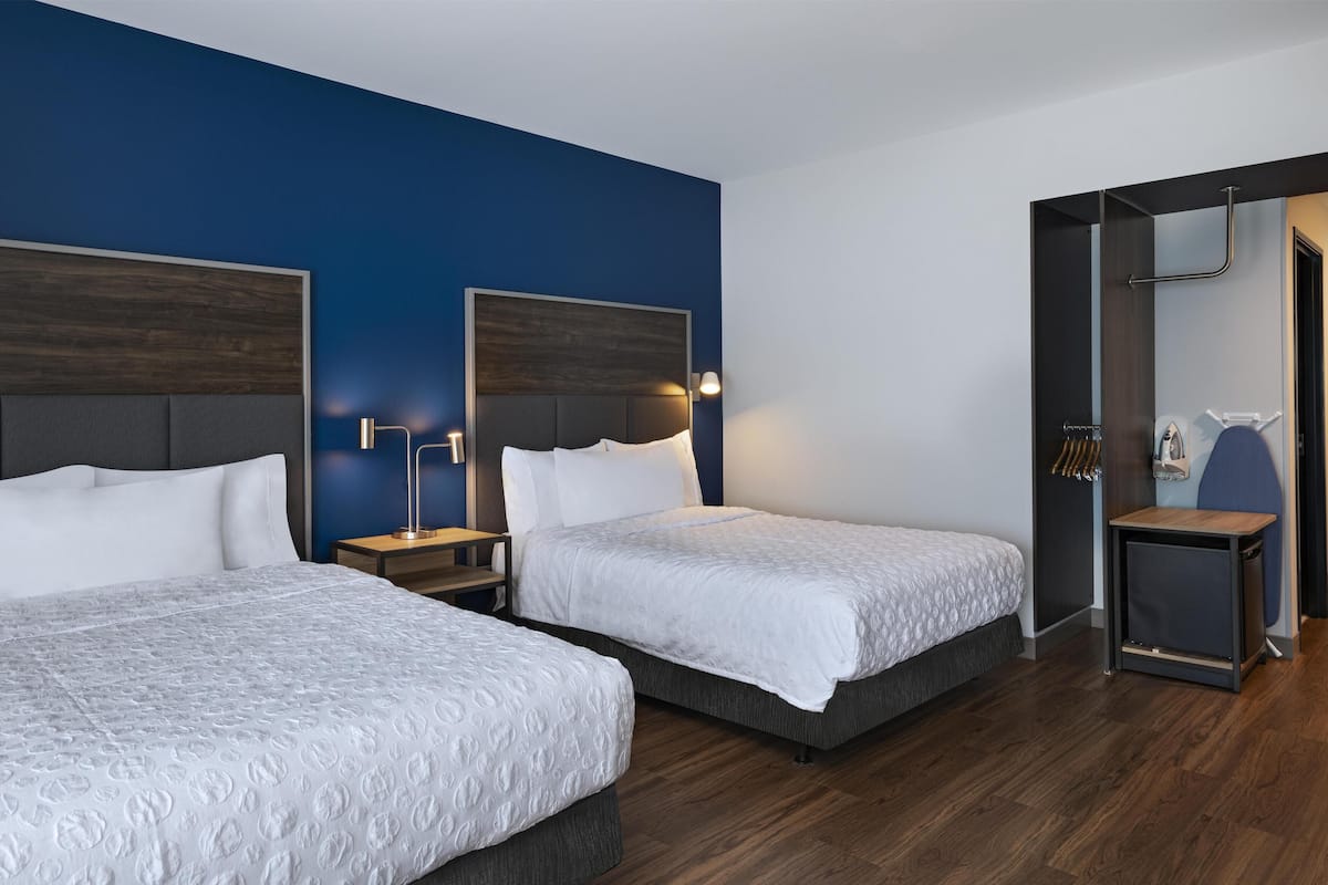 A hotel room with two beds, dark wooden headboards, white bedding, and a small bedside table with a lamp.