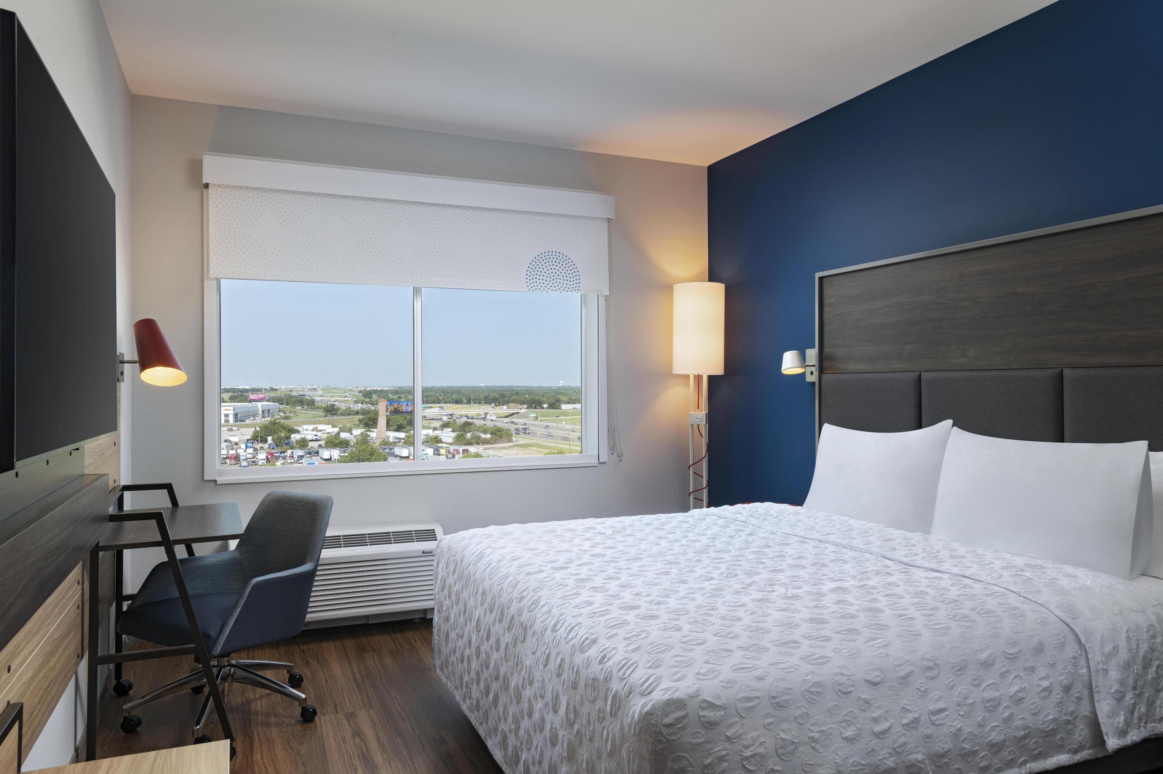 A hotel room with a large bed, a desk with a chair, a window with a view of the city, and a wall-mounted lamp.