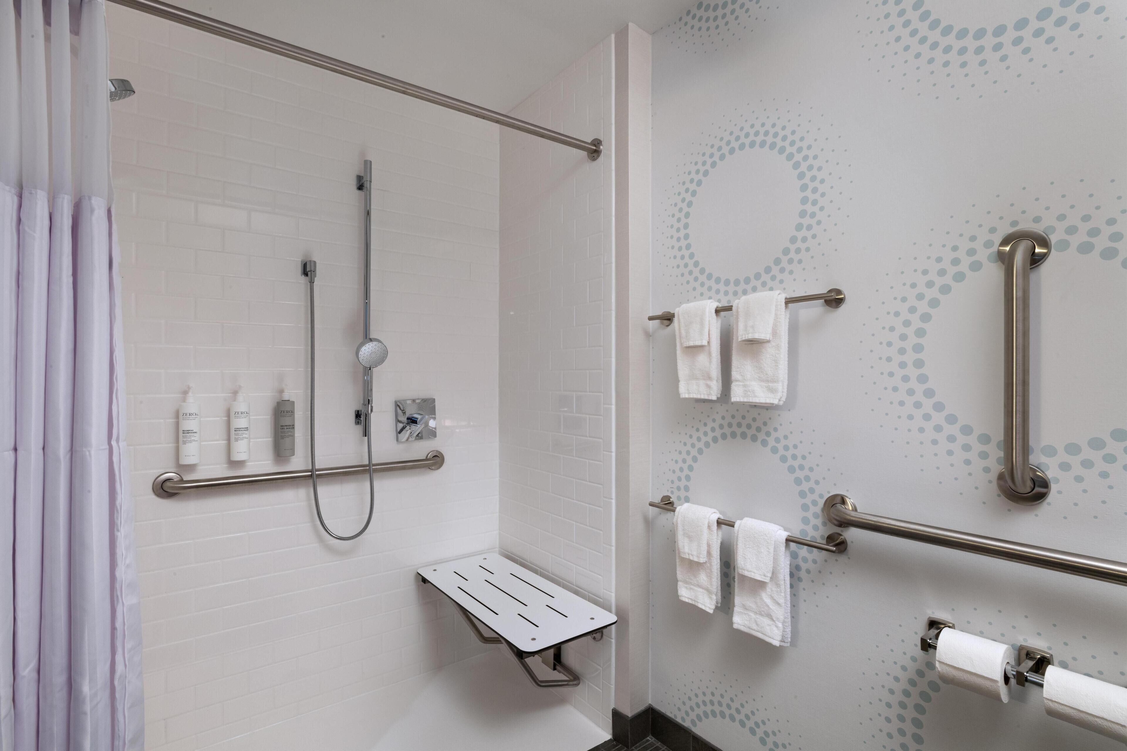 A modern bathroom with white tiled walls, a shower with a grab bar, a toilet with a grab bar, and a towel rack with multiple towels.