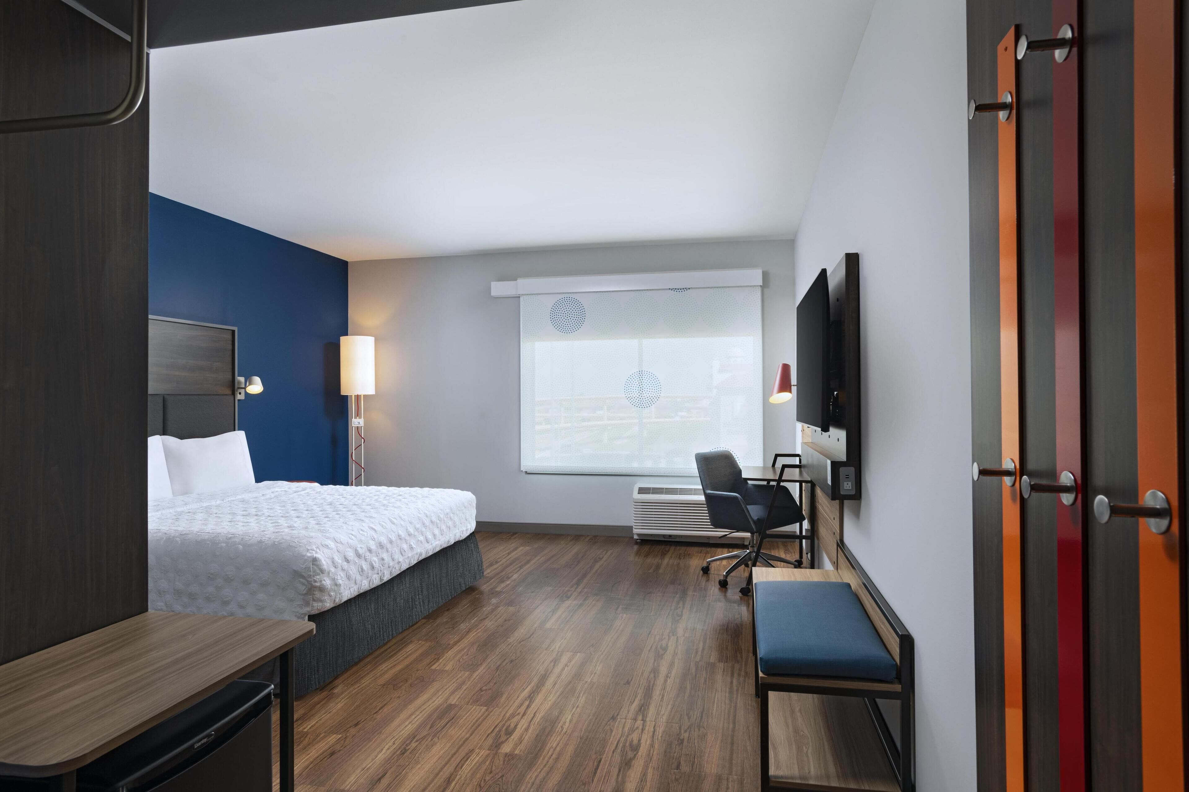 Ein Hotelzimmer mit Bett, Schreibtisch, Stuhl, Fernseher und einem Fenster mit Jalousien.