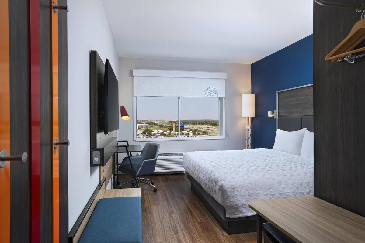 A hotel room with a bed, a desk, a chair, a TV, and a window with a city view.