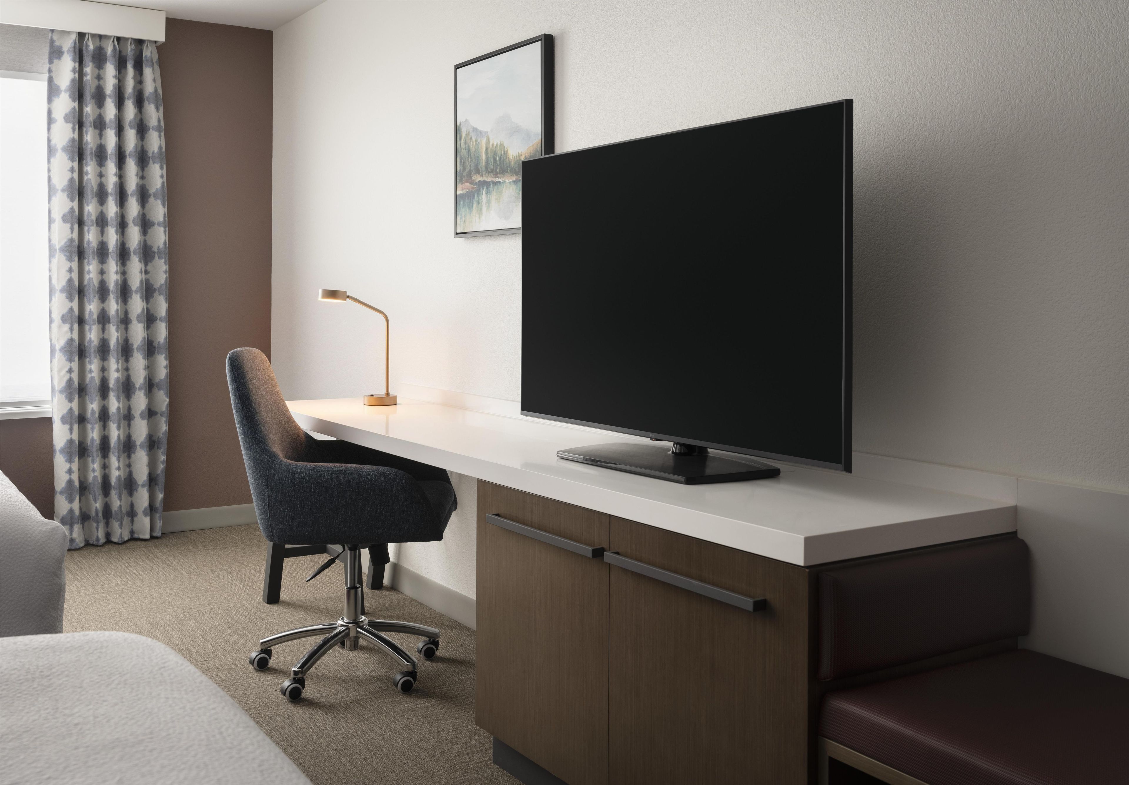 A hotel room with a bed, a desk with a computer monitor, a chair, and a lamp.