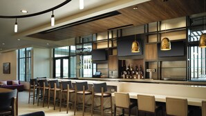 A modern bar area with wooden bar counters, multiple seating options including bar stools and tables, and a variety of bottles and glasses on the shelves. - Hilton Garden Inn Merced (Merced)