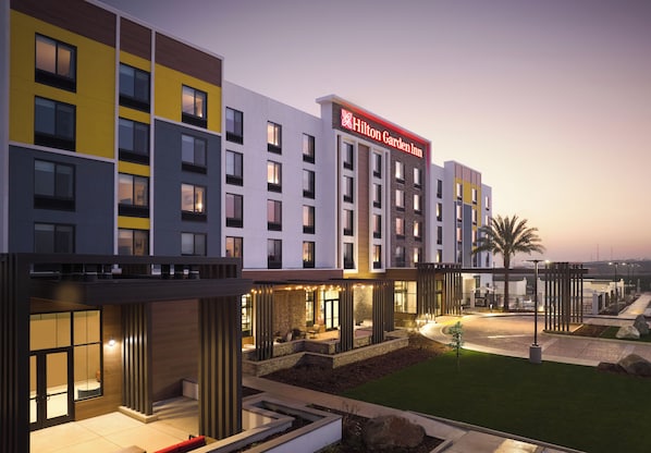 Un hotel de varios pisos con un llamativo letrero de Hilton Garden Inn, zonas de estar al aire libre y una entrada bien iluminada.