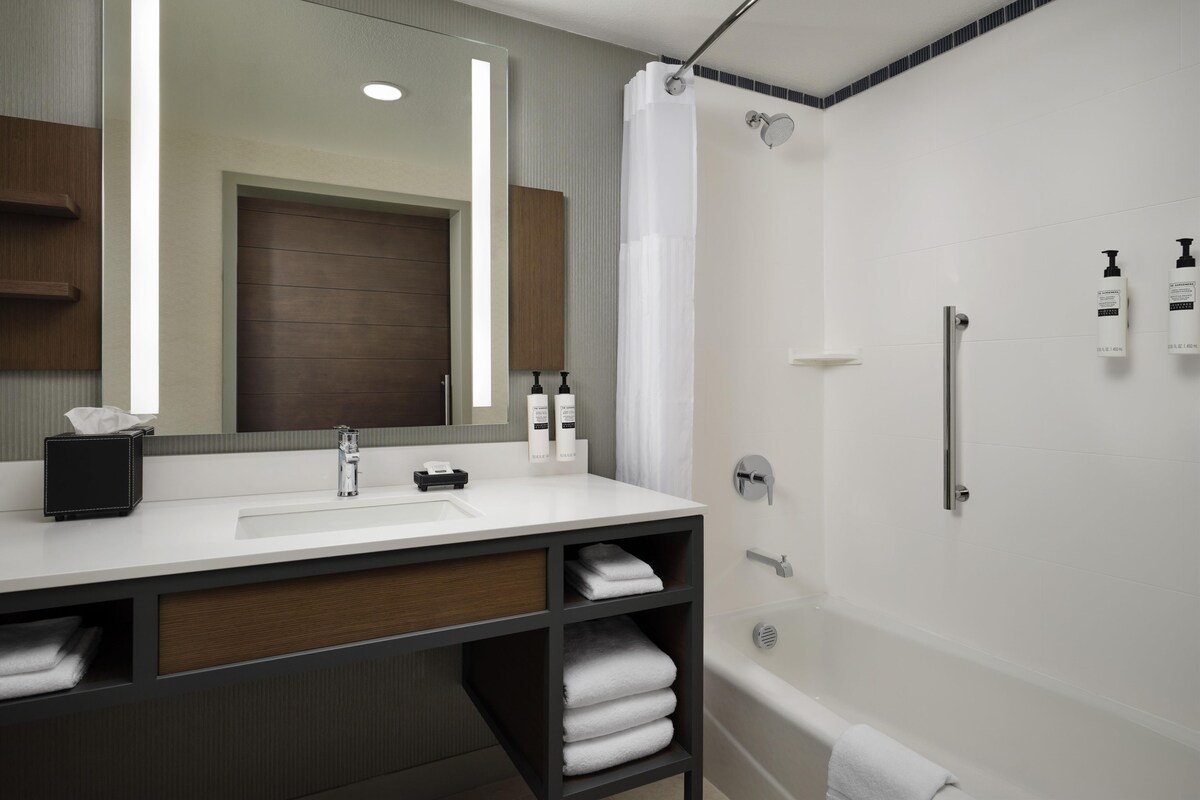 A modern bathroom with a white sink, mirror, and shower. There are towels and toiletries on the counter.