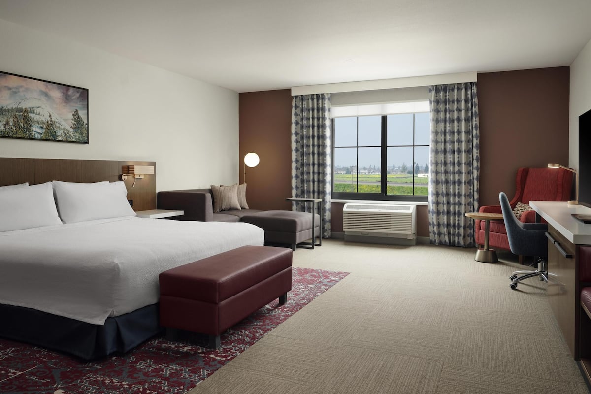 Une chambre d’hôtel avec un grand lit, un bureau, une chaise, un canapé, une fenêtre donnant sur un beau paysage et un tableau au mur.