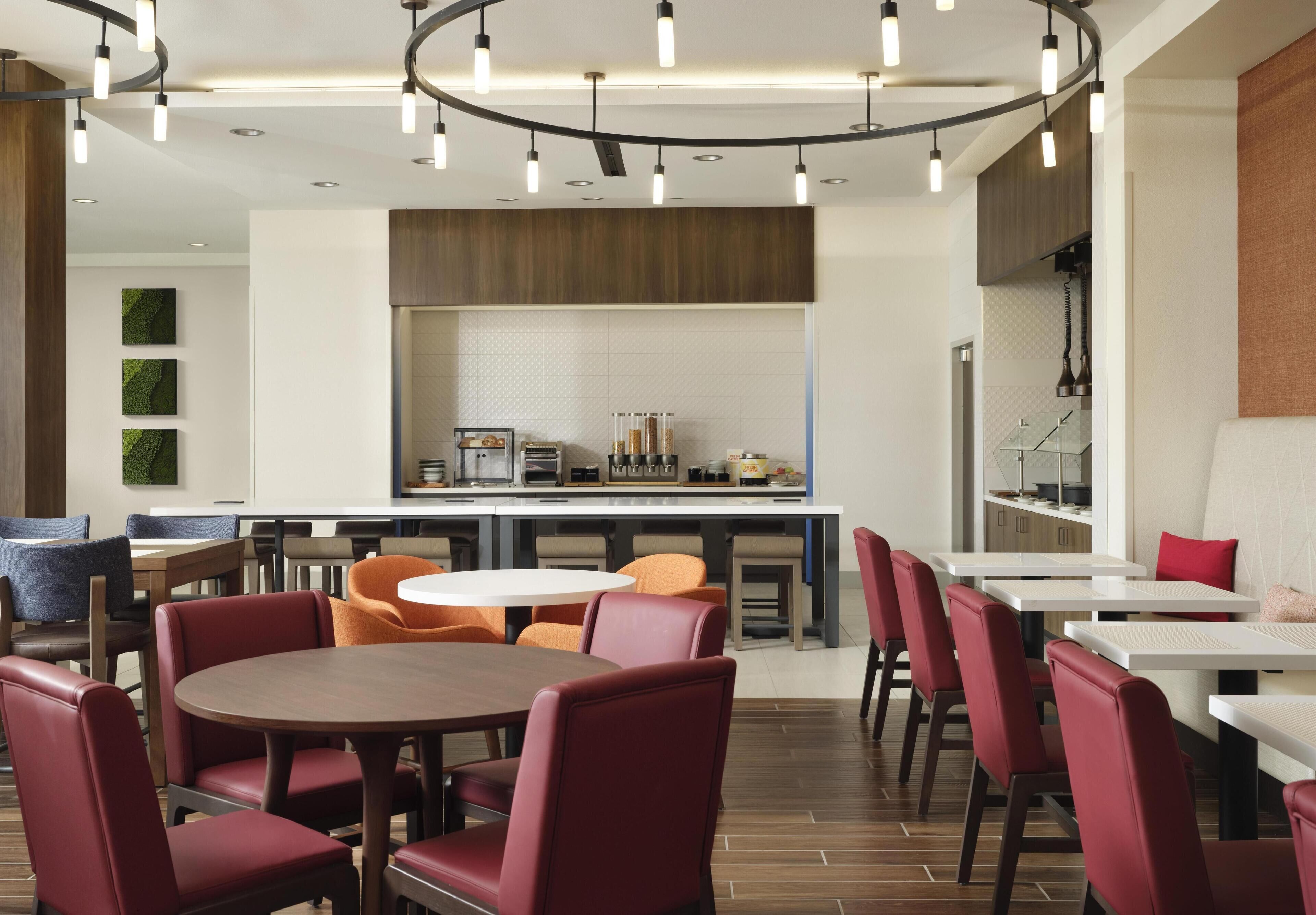 A modern restaurant with round tables, red and orange chairs, and a kitchen area in the background.