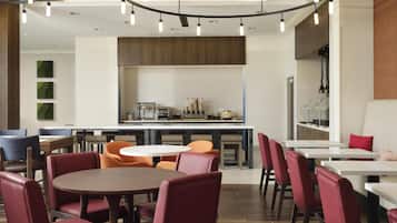 A modern restaurant with round tables, red and orange chairs, and a kitchen area in the background.