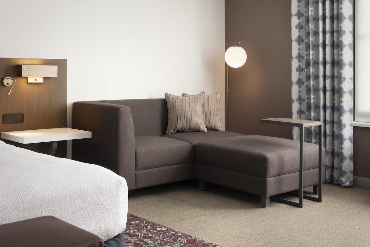 Une chambre d’hôtel moderne avec un canapé d’angle, une petite table, une lampe et une vue sur l’extérieur.