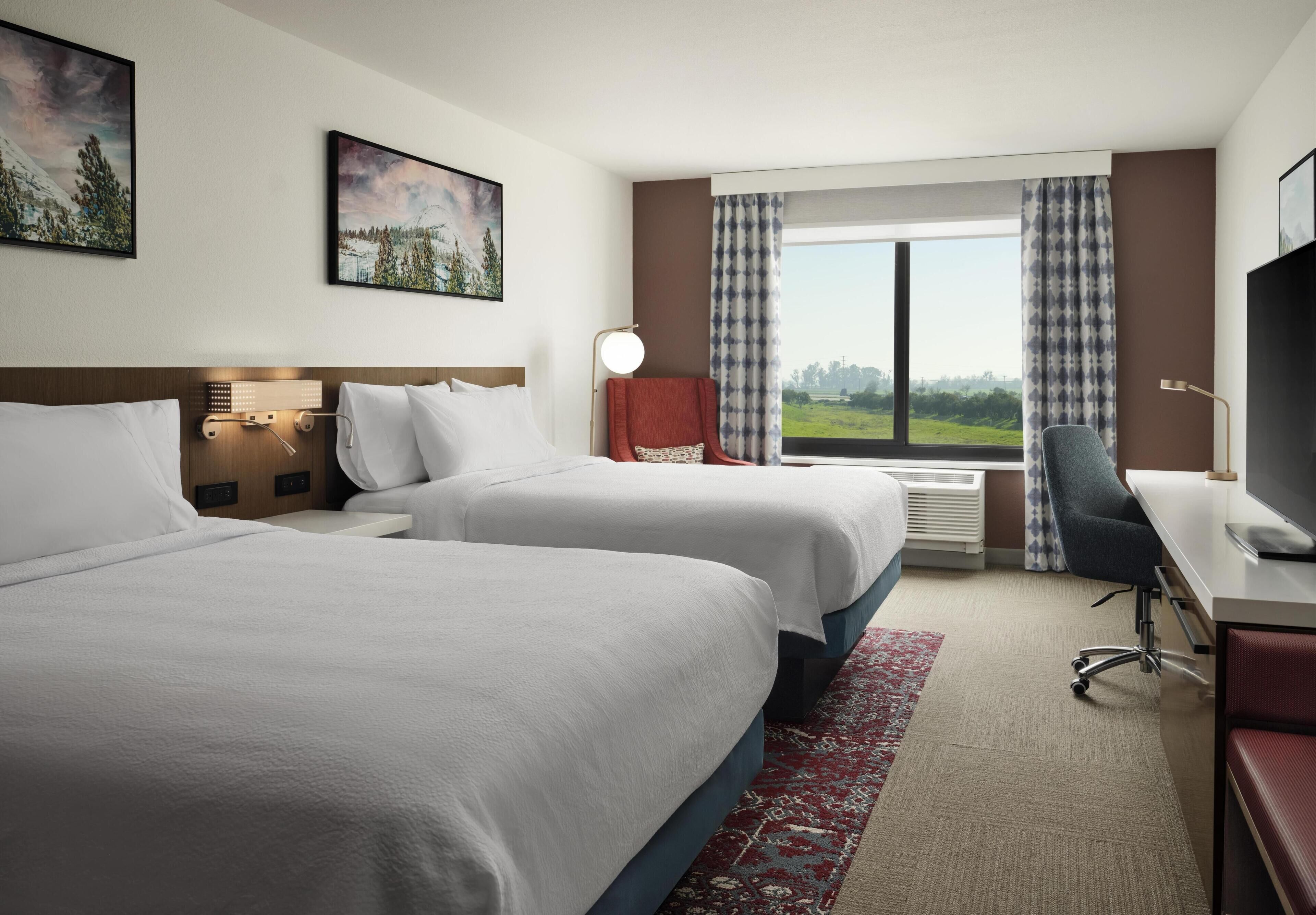Habitación de hotel con dos camas, un escritorio, una silla, un televisor y un ventanal con vistas.