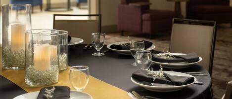 A dining table set with plates, cutlery, glasses, and candles, surrounded by chairs in a room with a carpeted floor and furniture in the background.