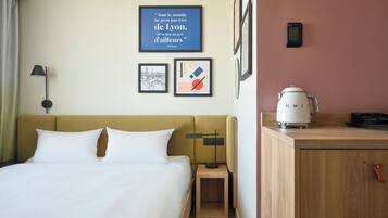 A hotel room with a bed, a nightstand, a lamp, a coffee maker, and a wall with framed pictures.