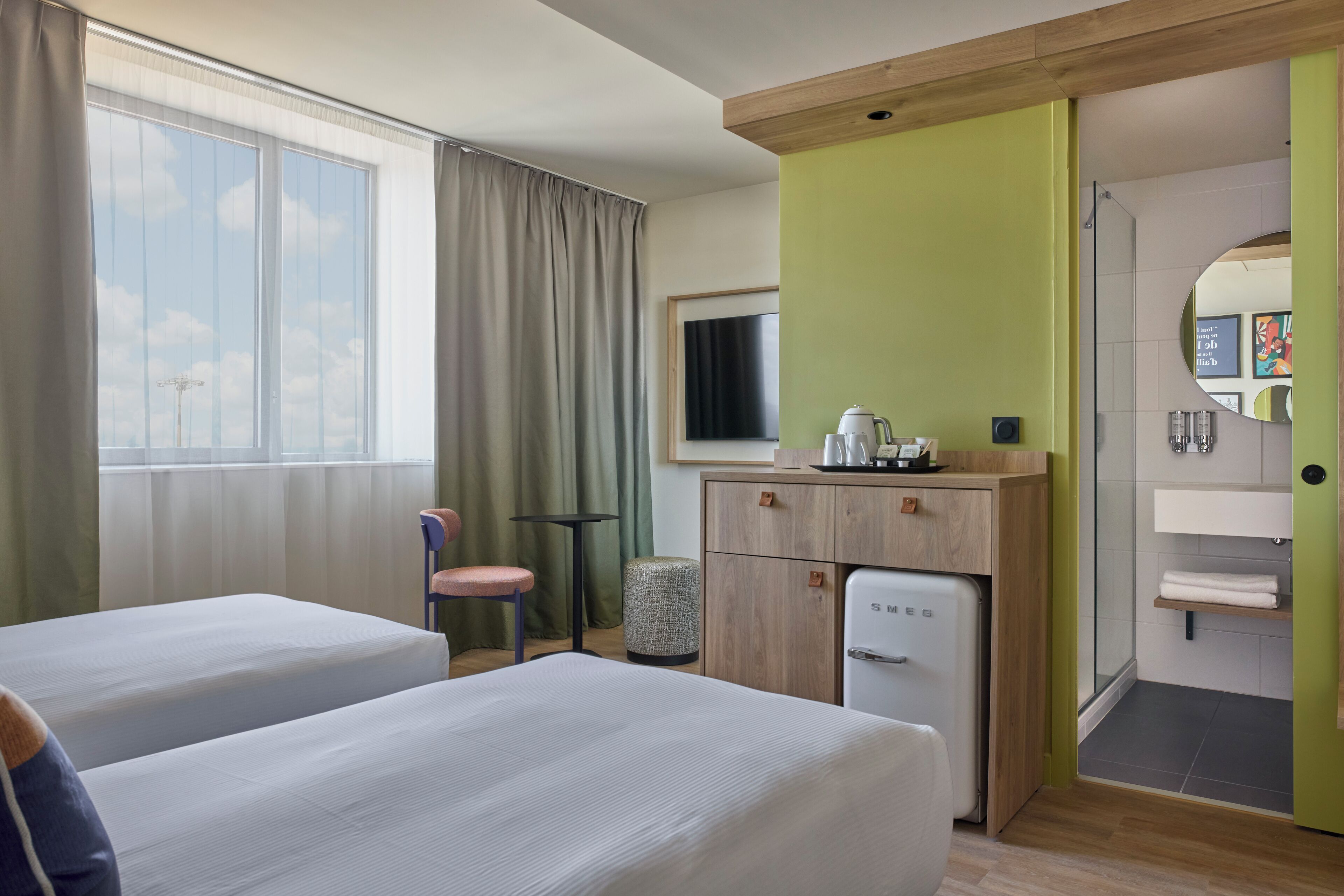 A hotel room with two beds, a small desk, a refrigerator, a chair, a window with curtains, and a view of the sky.