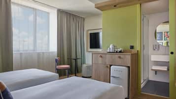 A hotel room with two beds, a small desk, a refrigerator, a chair, a window with curtains, and a view of the sky.