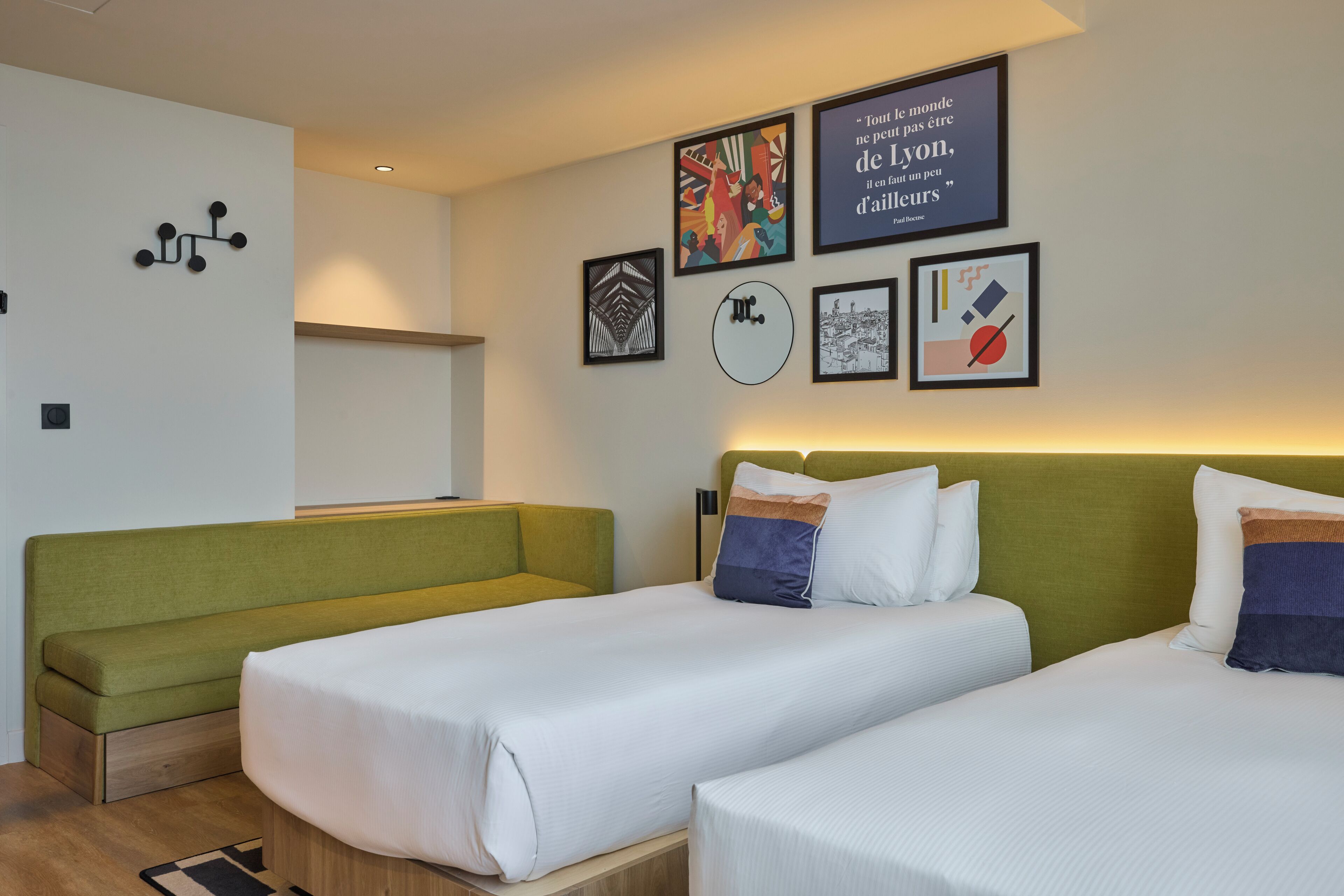 Une chambre d’hôtel avec deux lits simples, un canapé vert et un mur décoré de divers tableaux encadrés.