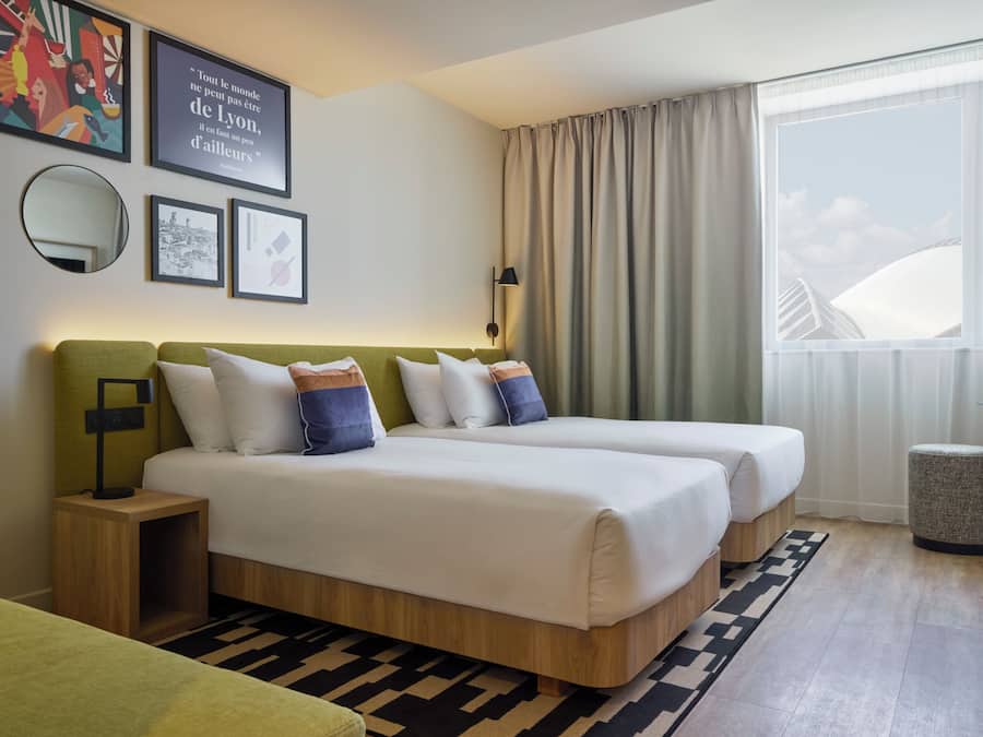 Una habitación de hotel con dos camas, cabecera verde, un ventanal con cortinas, un espejo redondo y cuadros enmarcados en la pared.