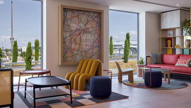 Een moderne woonkamer met een groot raam, een gele fauteuil, een rode bank en een boekenkast.