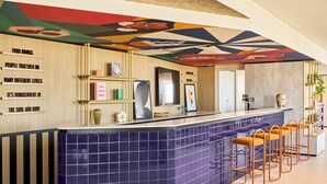 Een moderne bar met een blauw betegelde toonbank, houten planken en een kleurrijke muurschildering aan het plafond.