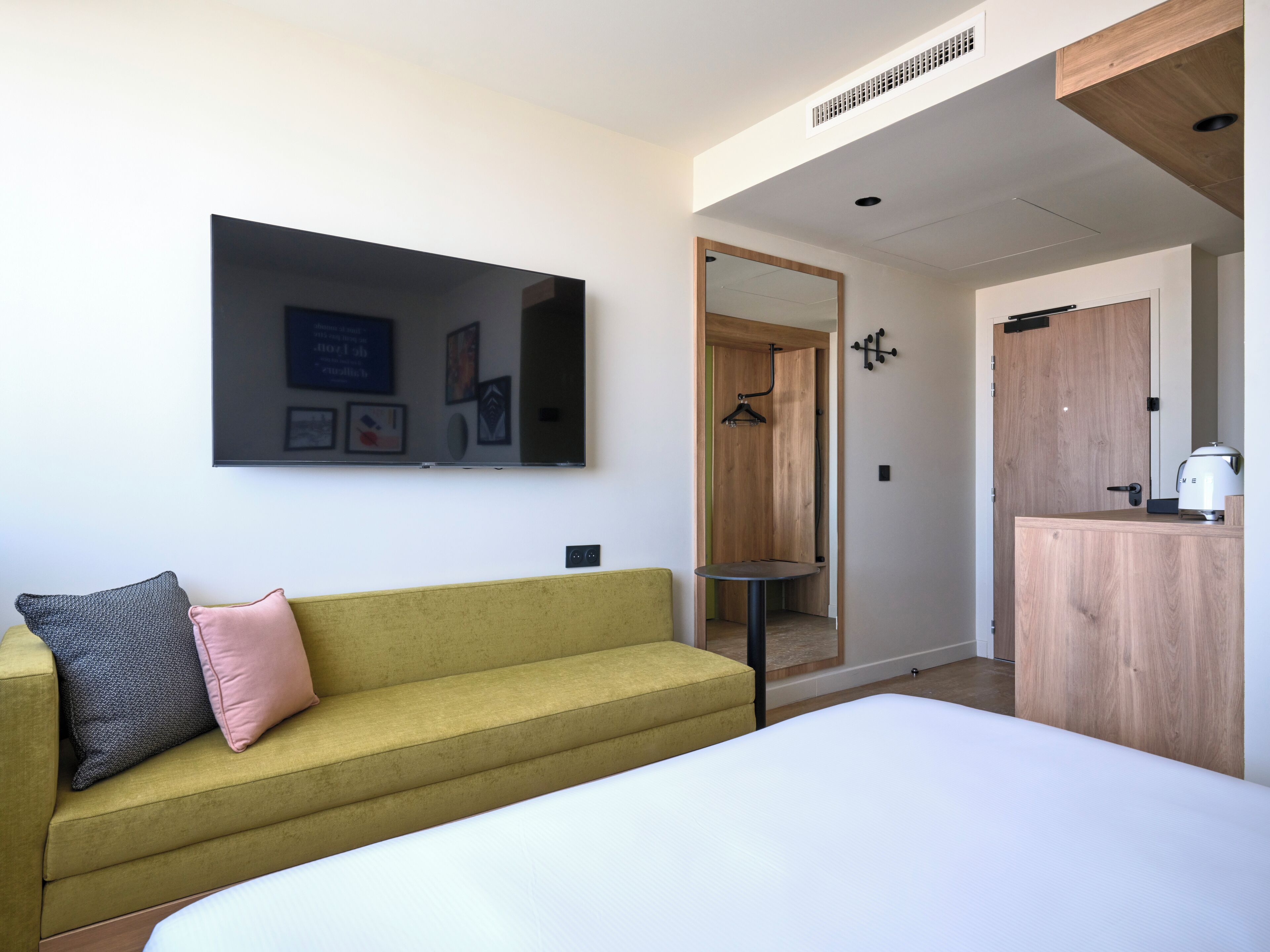 A modern hotel room with a large bed, a sofa, a small table, a mirror, and a wooden cabinet with a kettle.