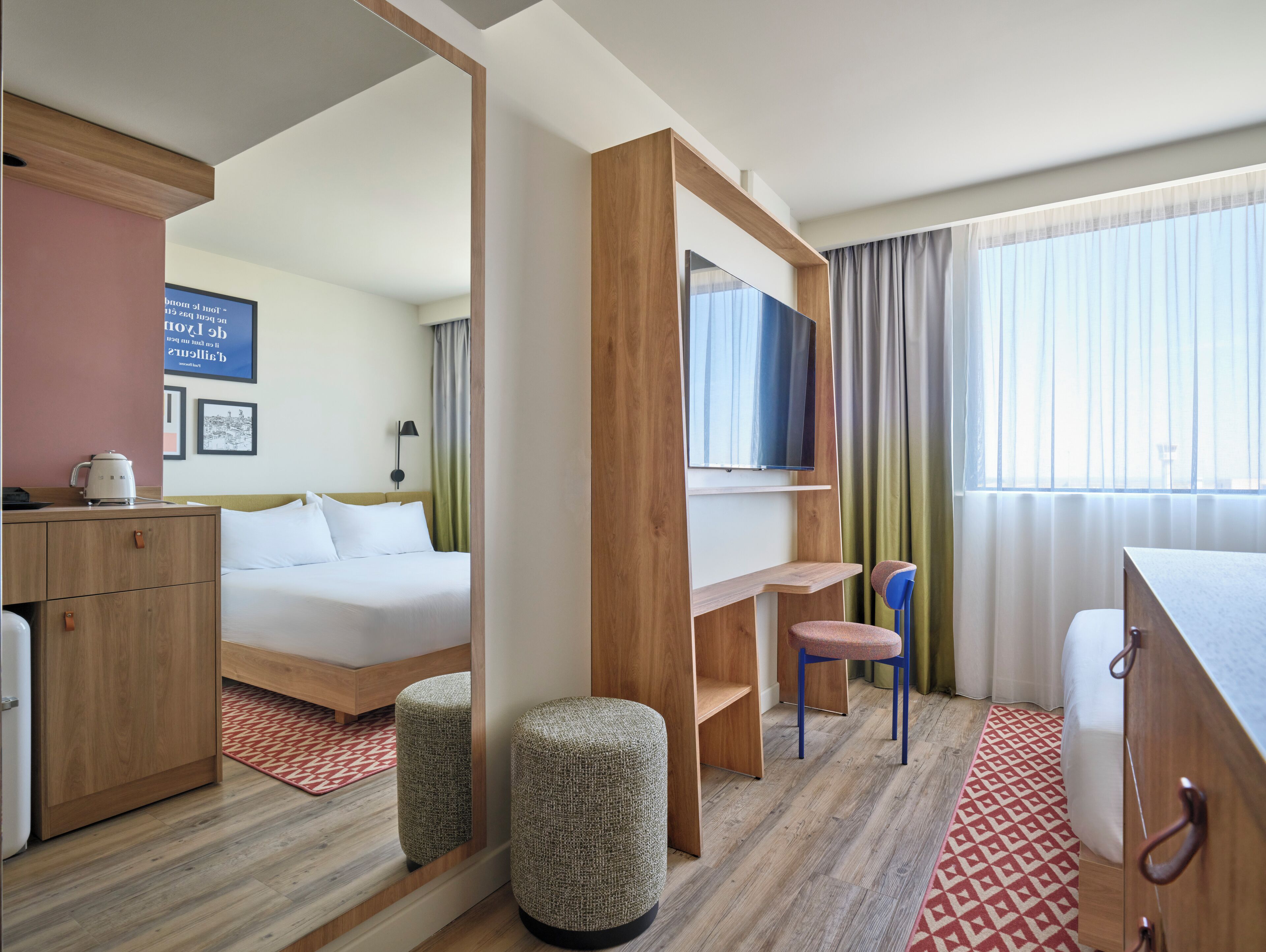 Une chambre d’hôtel comprenant un lit, un bureau, une chaise, une armoire, un miroir, une télévision et une fenêtre avec des rideaux.