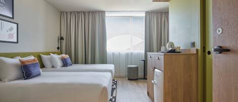 Habitación de hotel con dos camas, un escritorio, un cesto de basura pequeño y una ventana con cortinas.
