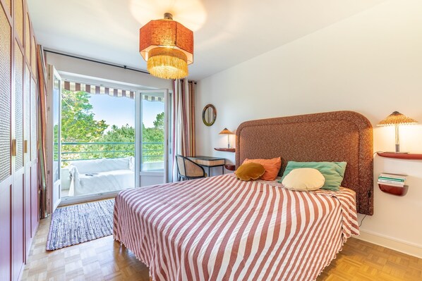 Un dormitorio con una cama grande, una mesita pequeña, una silla y vistas al exterior a través de una puerta corredera.
