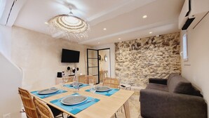 A dining area with a wooden table set for four, surrounded by wicker chairs. A stone wall and a gray sofa are visible in the background.