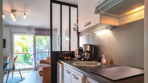 Cocina compacta con cafetera, lavaplatos y fregadero. Cuenta con una zona de comedor con una silla y mesa, además de un sofá. La cocina tiene una ventana con cortinas.