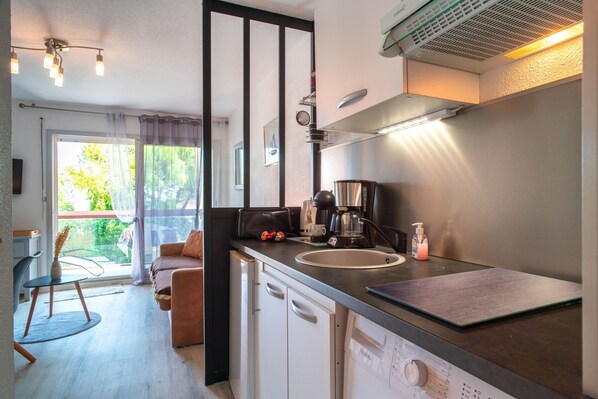 A compact kitchen with a coffee maker, a sink, and a dishwasher. There is a dining area with a chair and a table, and a sofa. The kitchen has a window with curtains. - Le Petit Saint Jacques - A quelques mètres de la mer (Sarzeau)