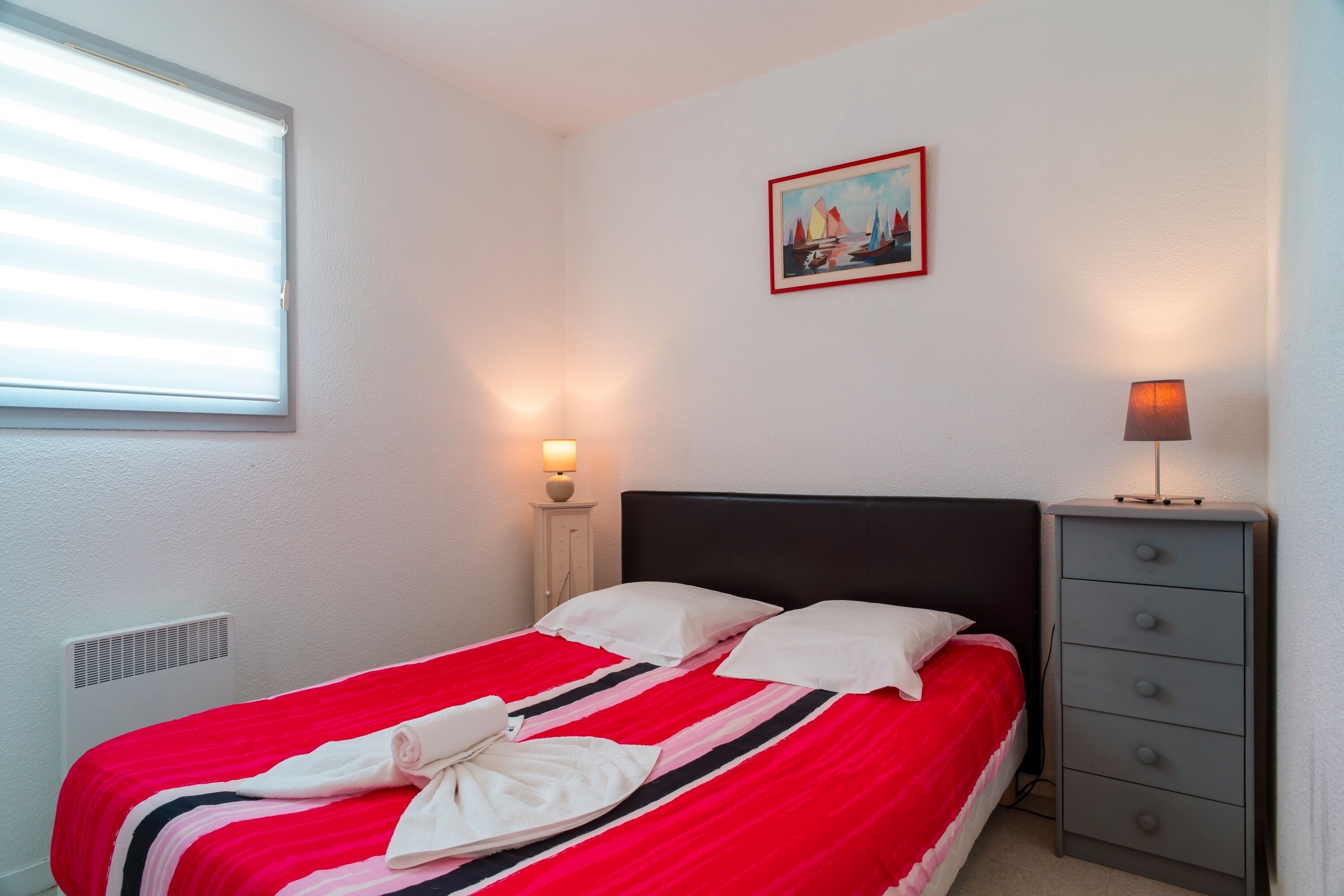 Un lit bien fait, recouvert d’une couverture rayée rouge et blanche, une petite table de chevet avec une lampe, et un tableau accroché au mur.