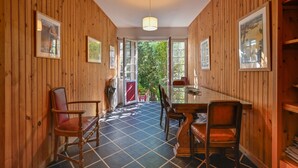 Ein Zimmer mit Holzwandverkleidung, einem Esstisch und Stühlen, gerahmten Bildern an den Wänden und Blick durch eine offene Tür ins Freie.
