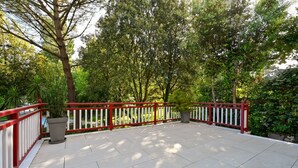 Ein überdachter Patio-Bereich mit roter und weißer Brüstung, umgeben von üppiger Bepflanzung und Bäumen.