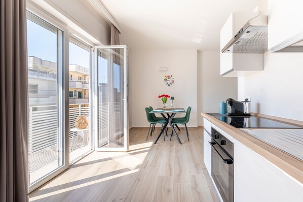Une cuisine moderne avec une porte vitrée donnant sur un balcon, un coin repas avec une table et des chaises, et une vue sur les bâtiments environnants.