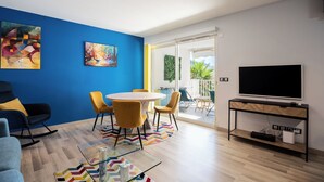 A modern living room with a blue accent wall, a round dining table with yellow chairs, a flat-screen TV on a wooden TV stand, and a glass coffee table.