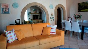 Una sala de estar con un sofá naranja, un comedor con sillas blancas, y una cocina visible a través de un arco.