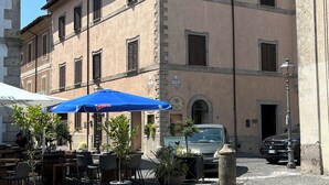 Un café al aire libre con mesas y sillas bajo un paraguas azul, un coche estacionado y un edificio con fachada de piedra y varias ventanas.