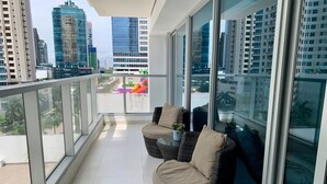 A balcony with two wicker chairs and a small table, large windows offering a city view.