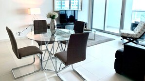 A modern living room with a glass table, four chairs, a sofa, a TV, and a large window with a city view.
