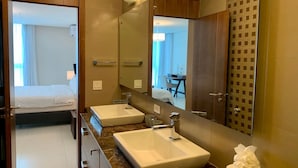 A modern bathroom with two sinks, a large mirror, and a bed visible through an open door.