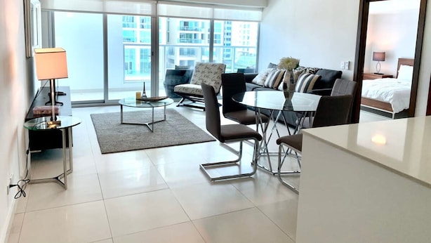 A modern hotel room with a large window, a sofa, a dining table, and a bed.