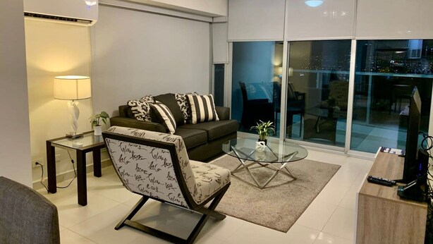 A modern living room with a sofa, a glass coffee table, and a chair. There is a lamp, a small table, and a TV stand. - Garden Cafe I Spacious 1 Bed Apt With Park View (Panama City)