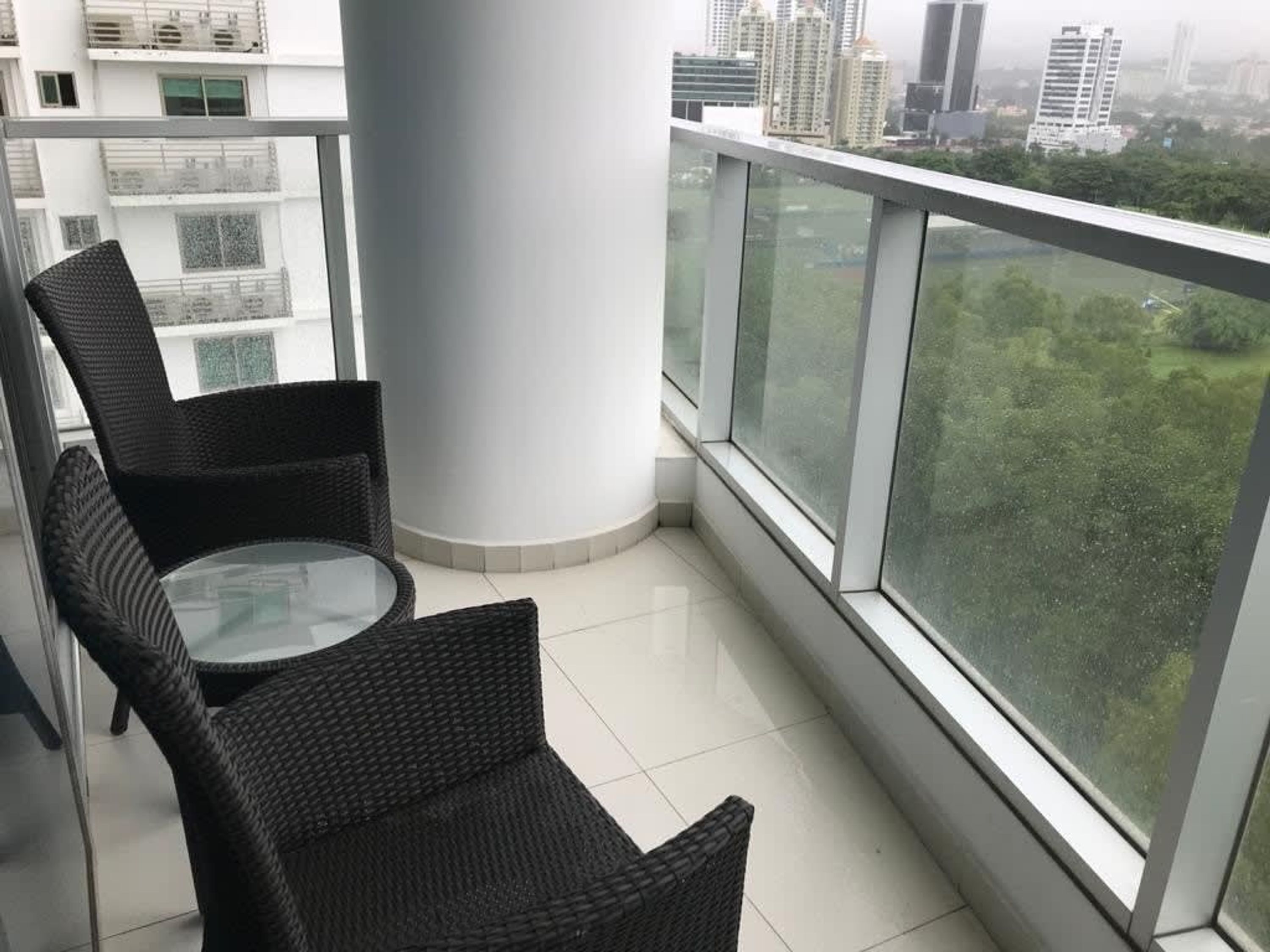 A balcony with wicker chairs and a glass table, overlooking a cityscape with high-rise buildings and greenery.