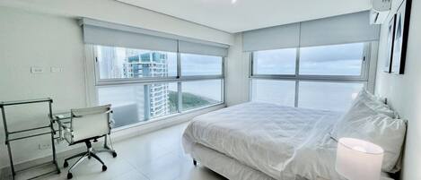 A modern hotel room with a large bed, a desk, and a chair. There are large windows providing a view of the city.