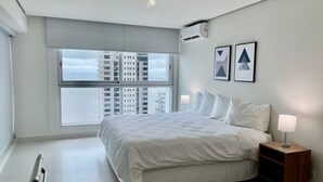 Une chambre moderne avec un grand lit, une table de chevet et une vue sur de hauts immeubles par une grande fenêtre.