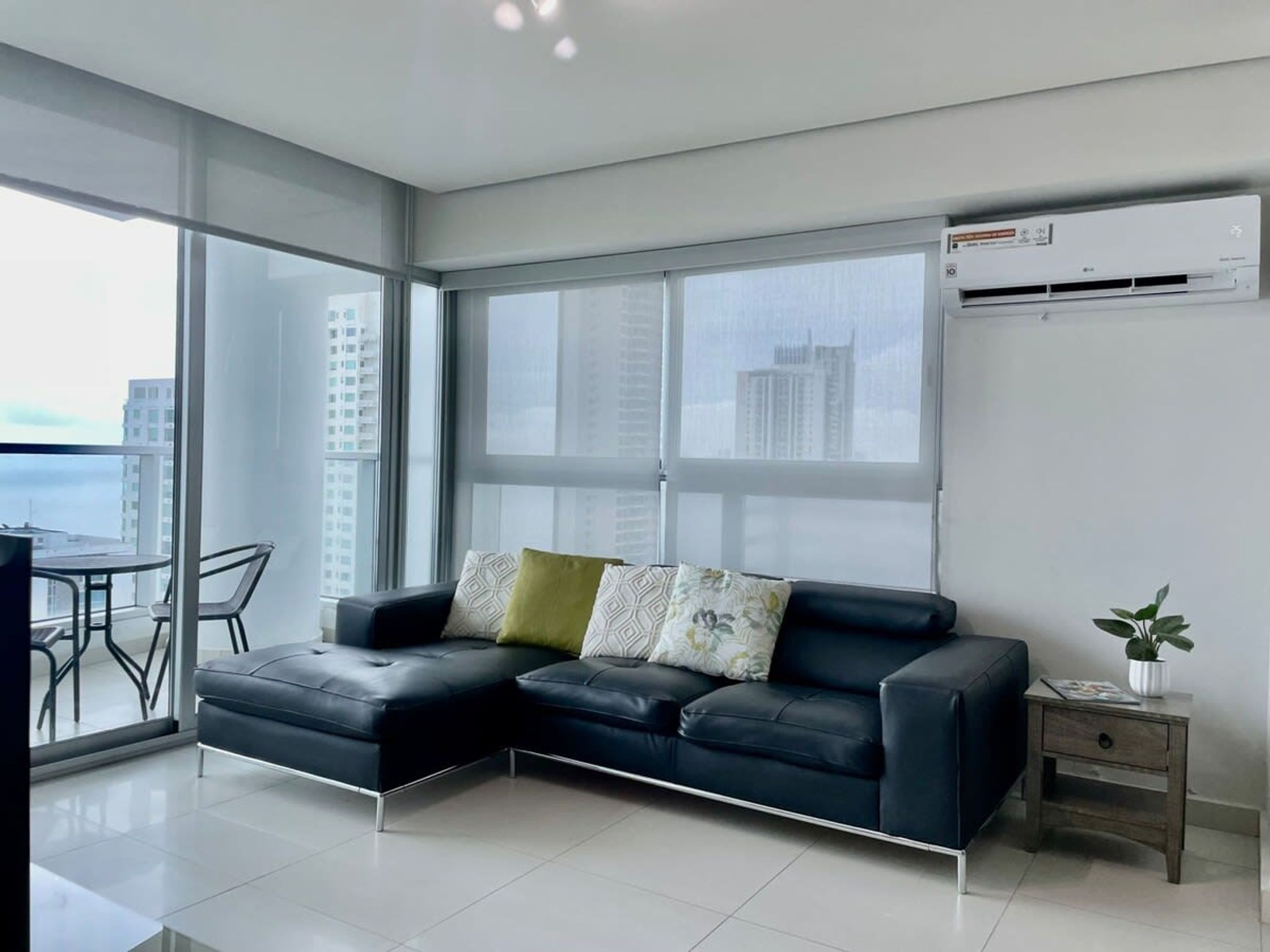 A modern living room with a black leather sofa, a small side table with a plant, and a view of the city through large windows.