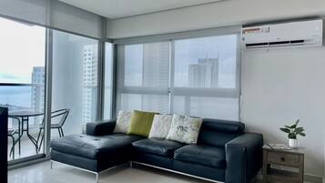 A modern living room with a black leather sofa, a small side table with a plant, and a view of the city through large windows.